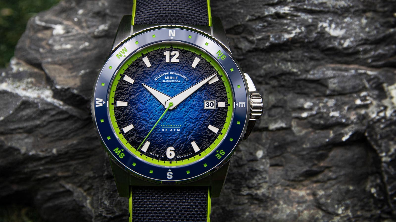Armbanduhr mit blauem Zifferblatt, grünem Rand und schwarzem Armband vor einem Felsen.