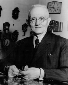 Ein älterer Mann mit Brille lächelt freundlich, umgeben von vielen Uhren an der Wand