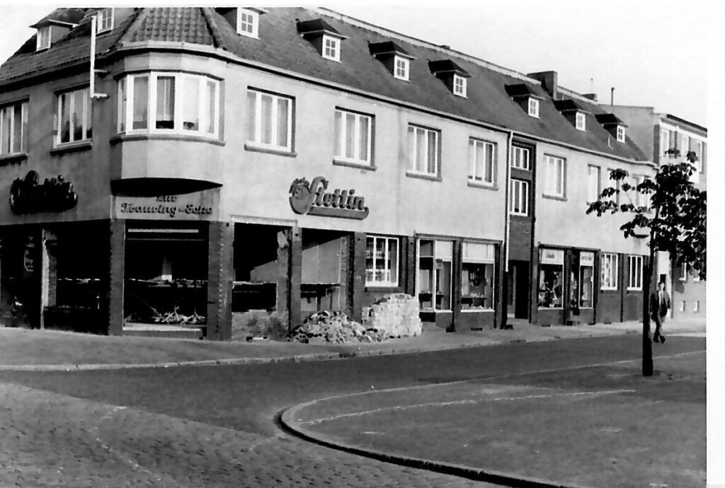 Ein grosses Eckgebäude mit mehreren Geschäften und Schaufenstern steht an einer gepflasterten Strasse, einige Baumreihen und Bauarbeiten sind sichtbar